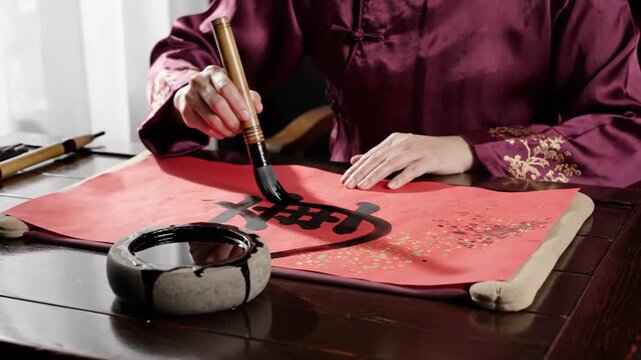 Person performing traditional Chinese calligraphy on red paper with ink brush
