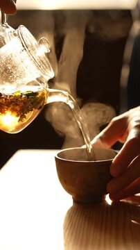 Hand pouring herbal tea from glass teapot into ceramic cup