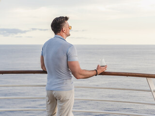 Stylish man with a glass of drink standing on the empty deck of a cruise ship against the backdrop of sea waves. Sunny day. Chill lifestyle, luxury travel. Perfect for holiday and travel themes © Svetlana