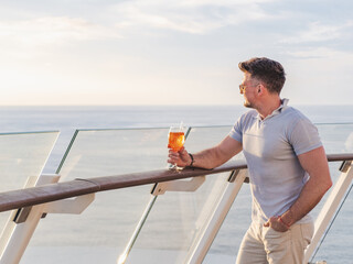 Fashionable man with a glass of drink standing on the empty deck of a cruise ship against the backdrop of sea waves. Sunny day. Chill lifestyle, luxury travel. Perfect for holiday and travel themes © Svetlana