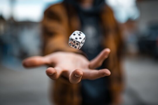 A man in a brown jacket tossing a silver dice into the air with his outstretched arm, a blurred background of a cityscape, and soft natural lighting