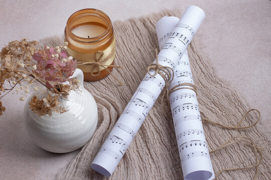 Aesthetic composition featuring music notes paper scrolls tied with twine, a burning candle in a glass jar, and hydrangea dried flowers on a textured neutral fabric background.