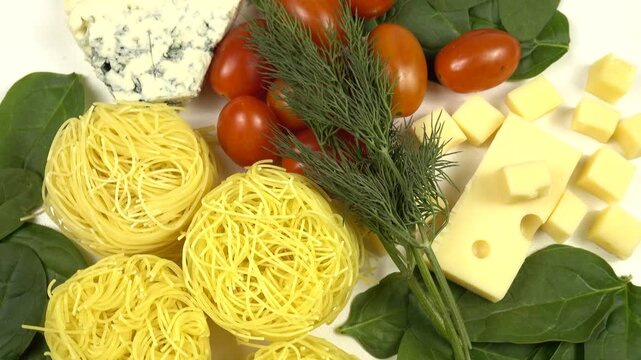 Dry Angel Hair pasta in nest-like shape, tomatoes, dill, various cheese and fresh spinach leaves, ingredients for making pasta, turning, top view