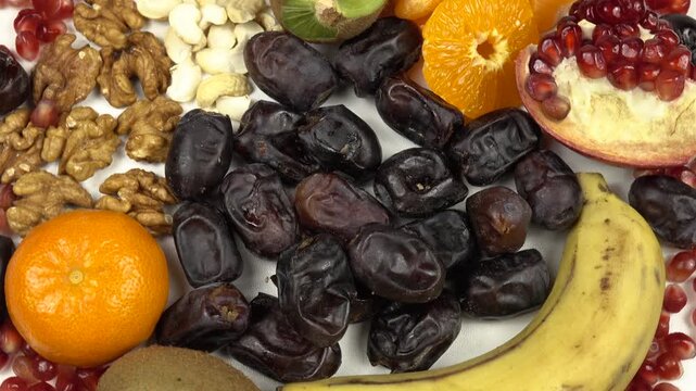 Tangerines, kiwi fruits, banana, pomegranate, black dates and nuts are on a white dish, top view, turning.