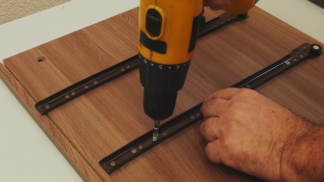 A furniture assembler screws drawer strips into place using a screwdriver. Medium shot.