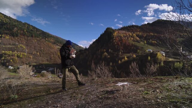 Man launching drone in mountains near valley, wearing beanie and backpack while stepping toward open ridge. Crisp autumn light, clouddappled sky, rugged slopes and distant village visible, conveying