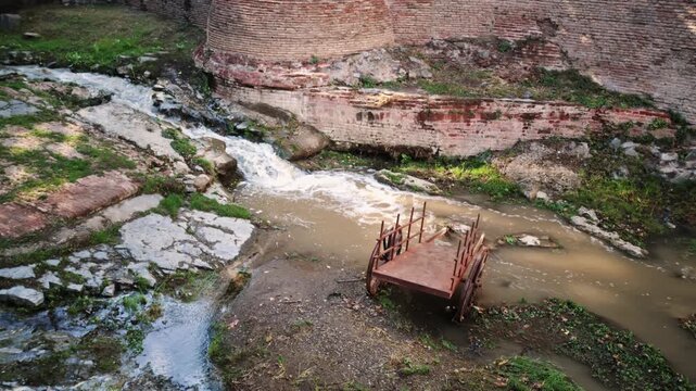 Rusted cart stranded in shallow creek, surrounded by eroded canyon walls, exposed bedrock moss and algae patches, silt lines tracing past water flow, muted sunlight casting warm highlights on corroded