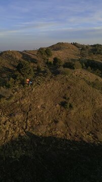 Vertical drone aerial of Mount Prau savanna landscape in Wonosobo, Central Java. Rolling grass hills glowing in sunrise light with mountain panorama background.