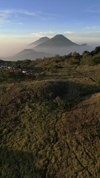 Vertical drone footage of colorful tents at Mount Prau campsite, Wonosobo. Savanna hills overlooking Mount Sindoro and Mount Sumbing under clear sky.