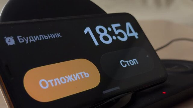 A smartphone sits on a black wireless charger, displaying a digital alarm clock with Russian text. Large snooze and stop buttons appear on the screen in an indoor setting.