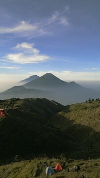 Vertical drone footage of Mount Prau savanna camp in Wonosobo. Colorful tents on rolling hills with Mount Sindoro and Mount Sumbing at sunrise.