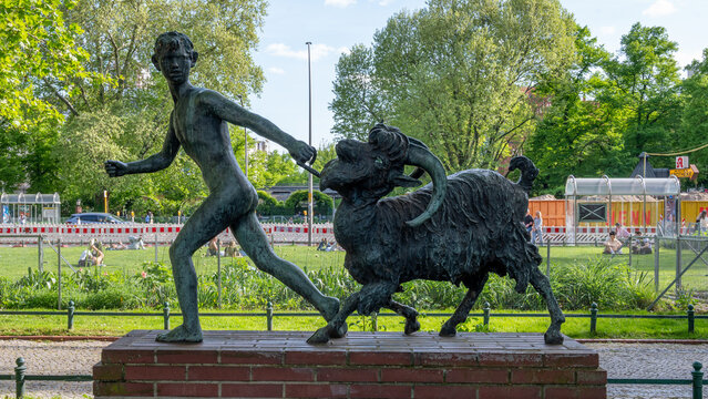 Bronze sculpture of a young shepherd leading a ram at Savignyplatz in Berlin. The artwork stands in a green urban park setting on a sunny day