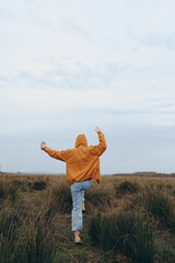 Fototapeta premium Woman lifestyle concept with a young woman wearing orange jacket and jeans walking through grassy field outdoors under cloudy sky. Casual fashion and nature exploration theme.