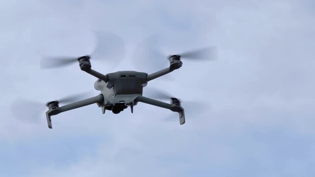 Close up of a quadcopter drone hovering in slow motion under a cloudy sky. Detailed view of the aircraft&acirc;s body and spinning rotors, set against a soft overcast background.