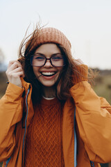 Smiling woman lifestyle wearing glasses, knit hat, and orange jacket outdoors in cool weather. Happy young female enjoying nature, casual fashion, and positive mood on autumn day.