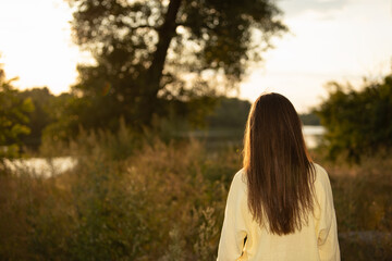 A woman in yellow looks out at the water during sunset. She stands in a field with trees and tall grass around her © Oleksandr Zinchenko