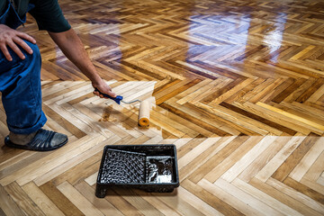 Professional worker varnishes oak parquet with a roller during floor renovation