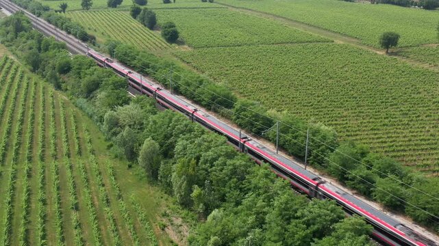 A sleek red high-speed train dashes through sprawling vineyards, contrasting nature and technology. Aerial drone view highlights its swift movement.