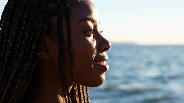 Young woman with braids enjoys calm ocean breeze at sunset. She smiles peacefully while relaxing by the water. Serenity and joy define this beautiful moment.