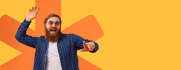 A man stands with a big smile, dancing in front of an orange and yellow background. He wears sunglasses and a plaid shirt. His arms are raised in a fun pose as he enjoys the moment. © Prostock-studio