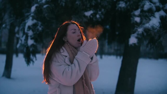 Young woman standing outdoors in snowy park during winter evening, breathing warm air into her hands and trying to keep warm in cold weather