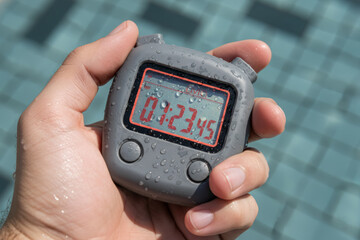 Wet male hand holding grey digital stopwatch with red numbers over blue swimming pool background representing race timing or sports training coaching concept