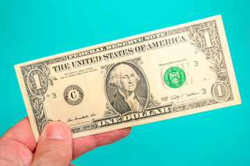 Close-up of a classic U.S. one-dollar bill series 2009 held between fingers against vibrant teal background.