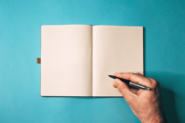 Overhead shot of open hardcover notebook with lined pages and black pen held above blank sheet on teal background, minimalist writing concept.