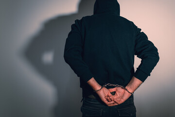 Rear view of a hooded person wearing handcuffs, face hidden in shadow, symbolizing detention, punishment, law enforcement, and accountability.