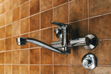 Wall-mounted chrome kitchen faucet against brown ceramic tiles, minimal interior design detail with clean reflective metal surface.