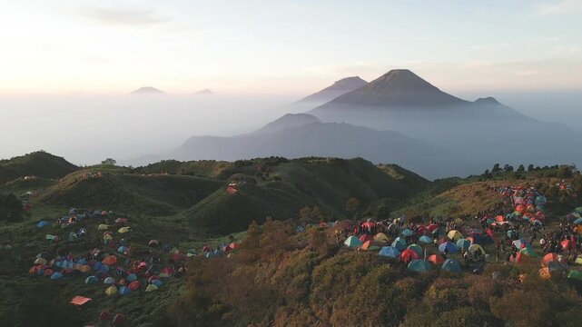 Drone view of Mount Prau campsite in Wonosobo, Central Java. Peaceful sunrise over savanna hills as hikers wake and enjoy serene views of Mount Sindoro and Sumbing.