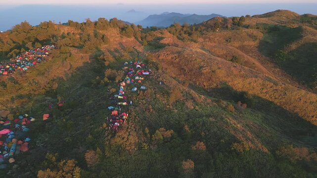 Golden sunrise light over Mount Prau savanna filled with colorful tents. Climbers and hikers gather in calm morning atmosphere with Sindoro and Sumbing on horizon.