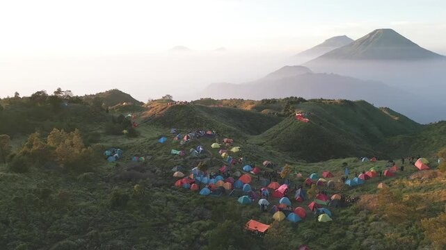 Wide drone shot of Mount Prau campsite surrounded by tranquil savanna. Peaceful hiking atmosphere with distant Sindoro and Sumbing mountains under soft sky.