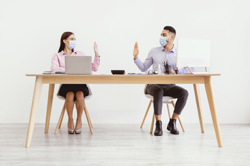 Obraz premium Two people sit at a wooden desk in an office setting. They are wearing masks and having a discussion. Each person has a laptop and is engaged in work.