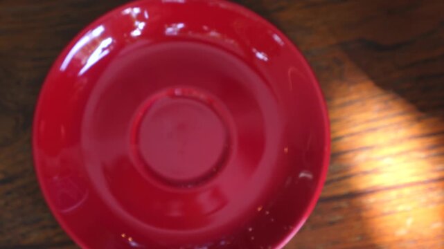 Top view of a woman's hands holding a red cup of cappuccino with beautiful latte art, she then picks it up and drinks the coffee, leaving an empty saucer on the wooden table
