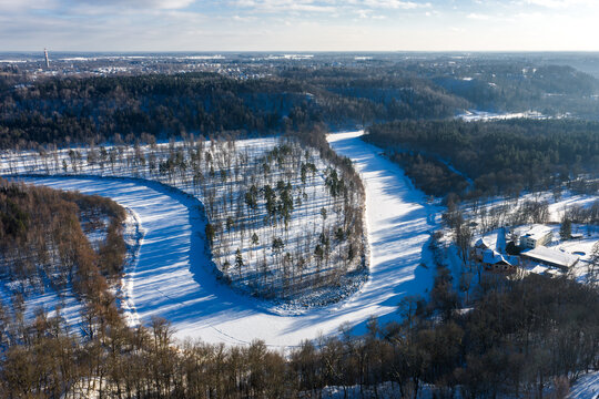 Aerial view of the Gauja River valley near Sigulda, Latvia, with a frozen oxbow, snowy peninsula, sparse pines and birches, and small snow covered buildings at right.