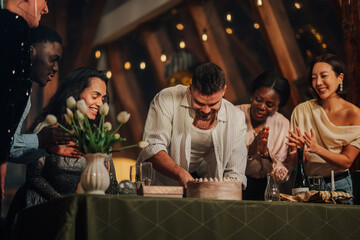 Diverse group of people enjoying a birthday celebration, smiling and clapping as a man cuts the...