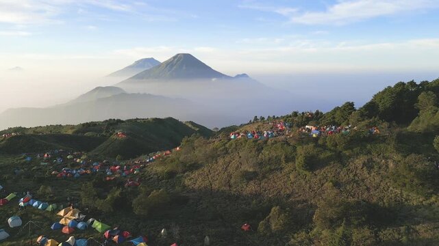 Drone view of serene savanna hills at Mount Prau. Hikers gather around tents, embracing fresh mountain air and majestic Sindoro Sumbing backdrop.