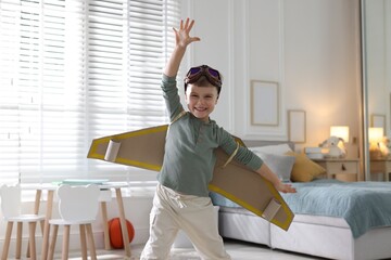 Portrait of smiling little boy with cardboard plane wings and goggles playing pilot at home
