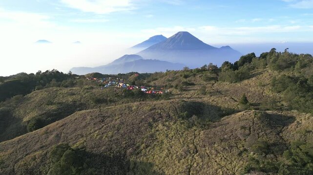 Beautiful aerial view of Mount Prau campsite at sunrise. Hikers resting in calm savanna landscape with Mount Sindoro and Mount Sumbing in background.