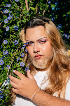 Man With Long Hair Poses Beside Vibrant Blue Flowers in Sunlight