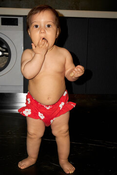 Little girl in red shorts stares at camera, hand in her mouth. 