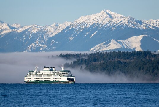 A Washington state ferry commute below the snow covered Olympic mountains. A foggy forest also seen in the background on a cold winter morning