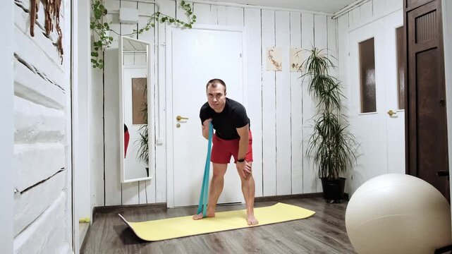 Caucasian man doing an exercise with an elastic band standing on a yoga mat at home interior. Pilates workout. Strengthening the muscles of the back, arms and shoulder girdle. High quality 4k footage.
