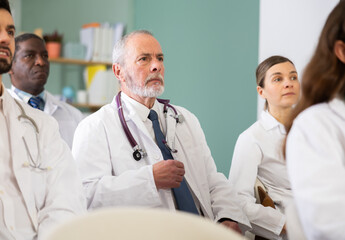Fototapeta premium Elderly doctor in a white coat against the background of doctors at a conference for advanced training, he listens attentively to the speaker