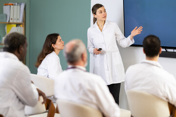 Obraz premium Adult female doctor in medical gown with remote control gives presentation to colleagues doctors in conference room