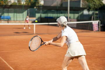 Senior woman in sportswear and a cap is playing tennis, she is holding a racket in her hands and making a ball pass