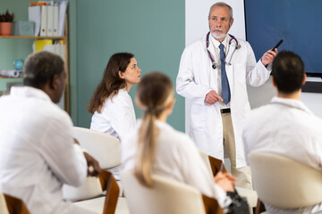 Fototapeta premium Mature doctor stands in front of a blackboard with a remote control in his hands, he gives a lecture to doctors for advanced training