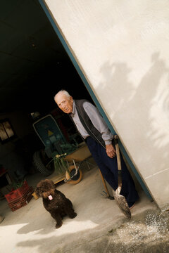 Elderly man stands at door with shovel and dog near farming area