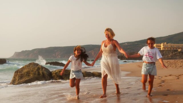 Happy mother holding hands and running with daughters along sandy beach, enjoying playful family bonding and joyful holiday by sea